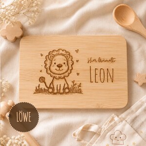May include: A rectangular light-colored wooden board featuring a laser-engraved illustration of a cartoon lion sitting in grass, surrounded by small hearts. The text "Hier brummelt" and the name "LEON" are engraved on the right side.
