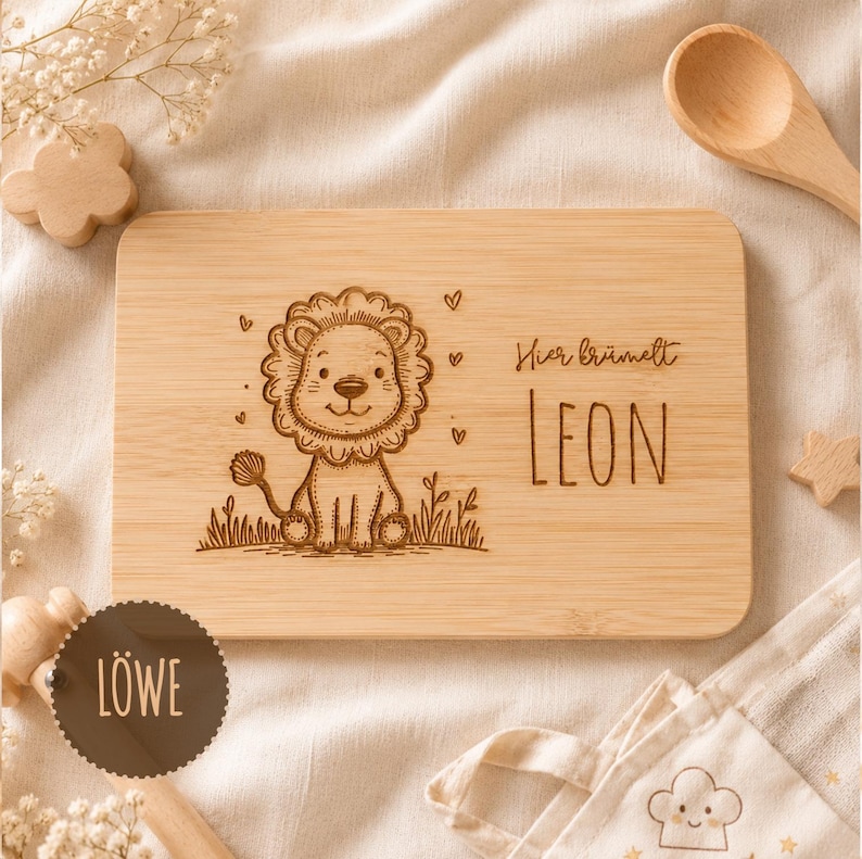 May include: A rectangular light-colored wooden board featuring a laser-engraved illustration of a cartoon lion sitting in grass, surrounded by small hearts. The text "Hier brummelt" and the name "LEON" are engraved on the right side.