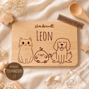 May include: A rectangular light brown wooden cutting board featuring a laser-engraved illustration of a cat, a small bird, and a dog. The text "Hier kr&uuml;melt" is engraved above the name "LEON" in a simple, dark brown sans-serif font.