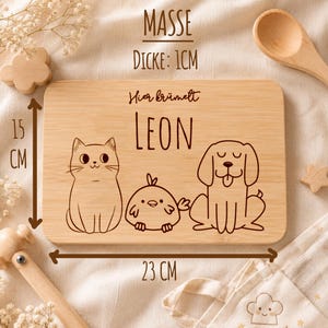 May include: A rectangular light brown wooden cutting board measuring 23 cm by 15 cm with a thickness of 1 cm. The board features laser-engraved illustrations of a cat, a small bird, and a dog, with the text "Hier kr&uuml;melt LEON" engraved above.