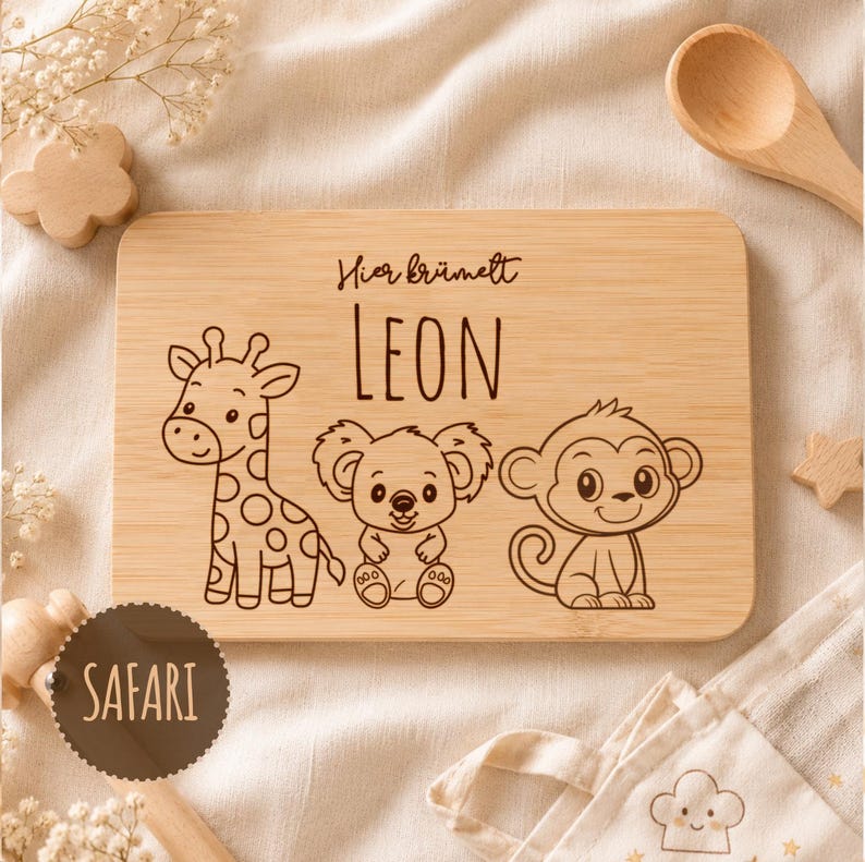 May include: A rectangular bamboo cutting board featuring a laser-engraved illustration of a giraffe, a koala, and a monkey. The text "Hier kr&uuml;melt" is engraved above the name "LEON" in a clean, dark font centered between the animal graphics.
