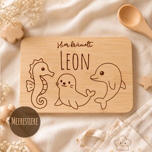 May include: A rectangular bamboo cutting board featuring a laser-engraved design of a seahorse, a seal, and a dolphin. The text "Hier kr&uuml;melt" is engraved above the name "LEON" in a simple, dark font, centered on the light brown wood surface.