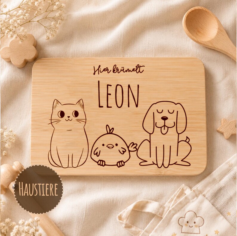 May include: A rectangular light brown bamboo cutting board featuring a laser-engraved illustration of a cat, a small bird, and a dog. The text "Hier kr&uuml;melt" is engraved above the name "LEON" in a simple, clean, dark brown font style.