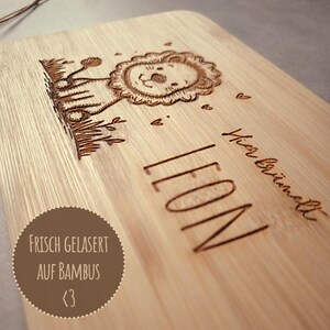 May include: A light-colored bamboo board featuring a laser-engraved illustration of a lion in tall grass, the name LEON, and the German text Hier br&uuml;llt, accompanied by a circular brown sticker with the text FRISCH GELASERT AUF BAMBUS <3.