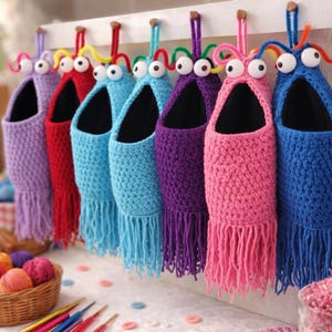 Op de afbeelding: Een rij kleurrijke, handgemaakte gehaakte wandzakken in verschillende kleuren, waaronder paars, rood, blauw en roze. Elke zak heeft franjes aan de onderkant en wiebelogen aan de bovenkant, wat een grillig ontwerp creëert. Ze hangen aan een wit houten bord.