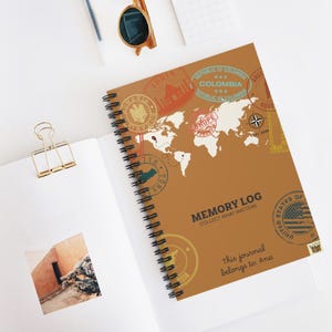 May include: A spiral-bound journal with a brown cover featuring a world map and travel-themed stamps. The text "MEMORY LOG COLLECT WHAT MATTERS" and "This journal belongs to: Ana" are also visible. Sunglasses and a paper clip are in the background.