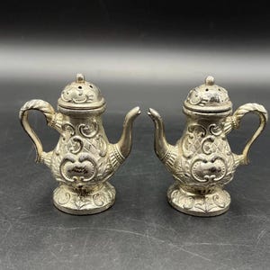 May include: Two silver-colored salt and pepper shakers in the shape of miniature teapots. Each shaker has a handle, spout, and a decorative lid with small holes. The shakers have an ornate, embossed design on the body and base.
