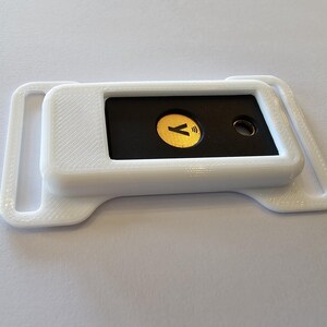 May include: A white, 3D-printed device with a black rectangular insert. The insert features a gold-colored circle with a 'Y' symbol and a wireless icon. The device has a rectangular shape with rounded edges and a loop on each side.