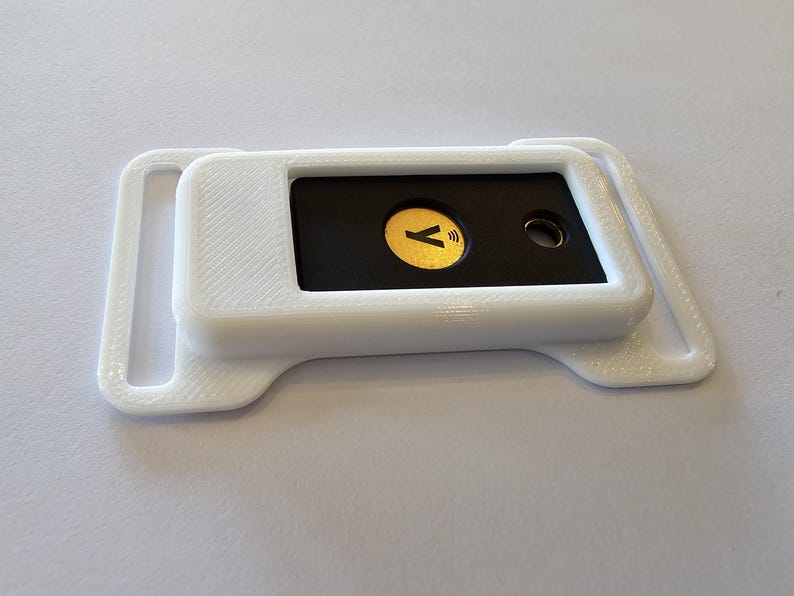 May include: A white, 3D-printed device with a black rectangular insert. The insert features a gold-colored circle with a 'Y' symbol and a wireless icon. The device has a rectangular shape with rounded edges and a loop on each side.