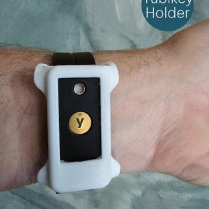May include: A white Yubikey holder with a black insert and a gold-colored button and logo. The holder is attached to a black strap. The text "Yubikey Holder" is in a teal circle in the upper right corner.
