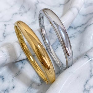 May include: Two shiny bangles, one gold and one silver, are displayed. The gold bangle has a warm, reflective surface, while the silver one gleams with a cool, polished finish. Both are smooth and rounded.