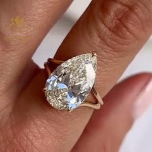 May include: A gold ring featuring a large, pear-shaped diamond. The diamond is the focal point, with a brilliant cut that reflects light. Another ring with rectangular diamonds is visible in the background. The ring is worn on a finger.