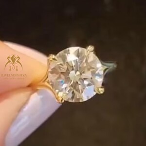 Puede incluir: Un anillo de oro con un gran diamante de talla redonda. El diamante está sujeto por cuatro garras. El anillo se muestra sobre un fondo oscuro.