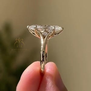May include: A gold engagement ring with a large, oval-cut diamond. The ring's band is a polished gold, and the diamond is held in place by delicate prongs. The ring is held up against a blurred background.