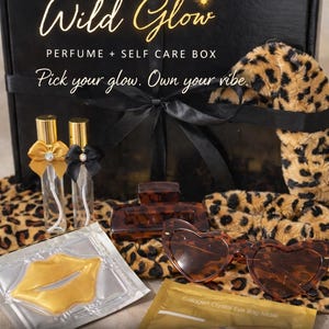 May include: A black gift box labeled "Wild Glow" with perfume and self-care items. The box includes perfume bottles, lip masks, eye masks, hair clips, and heart-shaped sunglasses. The box is tied with a black ribbon and sits on a leopard print fabric.