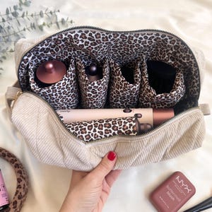 May include: Beige corduroy cosmetic bag with a leopard print interior. The bag is open, revealing compartments for hair styling tools, including a Dyson hair dryer. A hand with red nail polish holds the bag.