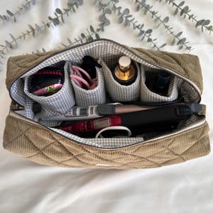 May include: A quilted corduroy cosmetic bag in a light brown color, open to reveal multiple compartments. The bag contains various beauty products, including bottles, hair tools, and accessories. The interior features a gray and white gingham pattern.