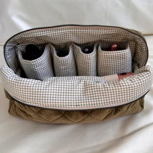 May include: Open olive green quilted corduroy cosmetic bag with a gingham interior. The bag has a zipper closure and multiple interior pockets holding makeup items. The bag is designed to organize and store cosmetics.