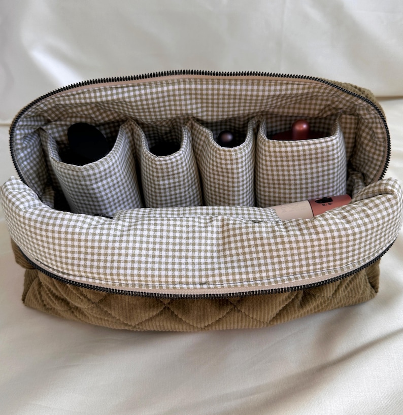 May include: Open olive green quilted corduroy cosmetic bag with a gingham interior. The bag has a zipper closure and multiple interior pockets holding makeup items. The bag is designed to organize and store cosmetics.