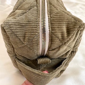 May include: Olive green corduroy bag with a quilted pattern and a gold zipper. The bag has a rectangular shape with a small pocket on the side. The corduroy fabric has a ribbed texture.