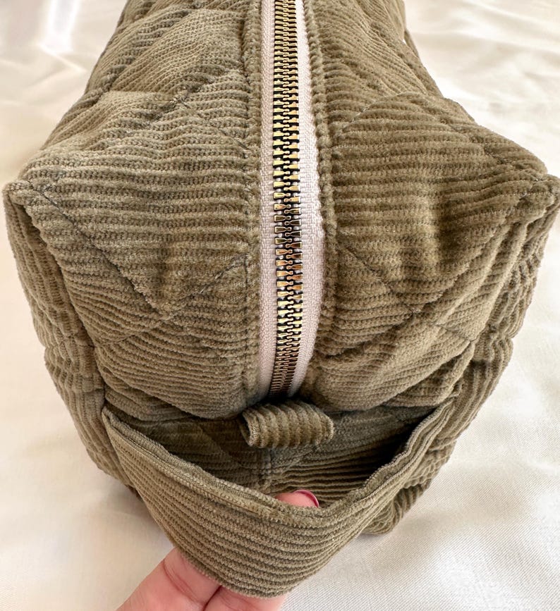 May include: Olive green corduroy bag with a quilted pattern and a gold zipper. The bag has a rectangular shape with a small pocket on the side. The corduroy fabric has a ribbed texture.