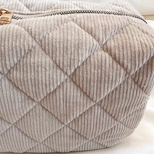 Peut inclure: Gros plan sur une trousse de maquillage en velours c&ocirc;tel&eacute; beige avec un motif de losanges matelass&eacute;s. Le sac est dot&eacute; d'une fermeture &eacute;clair dor&eacute;e et d'une forme arrondie. Le tissu en velours c&ocirc;tel&eacute; a une texture c&ocirc;tel&eacute;e.