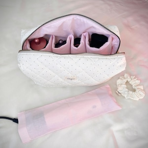 May include: A white quilted cosmetic bag with small black dots and a pink interior featuring multiple compartments holding hair styling attachments. A matching pink fabric sleeve and a white polka dot scrunchie are included. The label reads Elvyva.