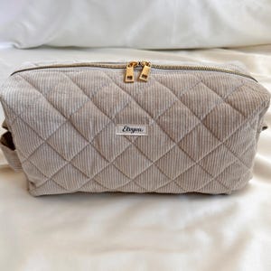 Peut inclure: Une trousse de maquillage en velours c&ocirc;tel&eacute; beige clair avec un motif losange matelass&eacute;. Le sac a une fermeture &eacute;clair dor&eacute;e et une petite &eacute;tiquette qui dit "Elvyva". Le sac est rectangulaire.