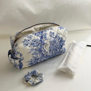 May include: A quilted toile de jouy cosmetic bag in white and blue with a zipper closure and a small label reading "Eleyva". The set includes a matching fabric scrunchie and a long, rectangular white fabric pouch with a black drawstring.