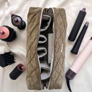 May include: A corduroy cosmetic bag in olive green, open to reveal a gingham lining and interior pockets. Various hair styling tools, including a hair dryer, curling wands, and brushes, are arranged around the bag on a white surface.