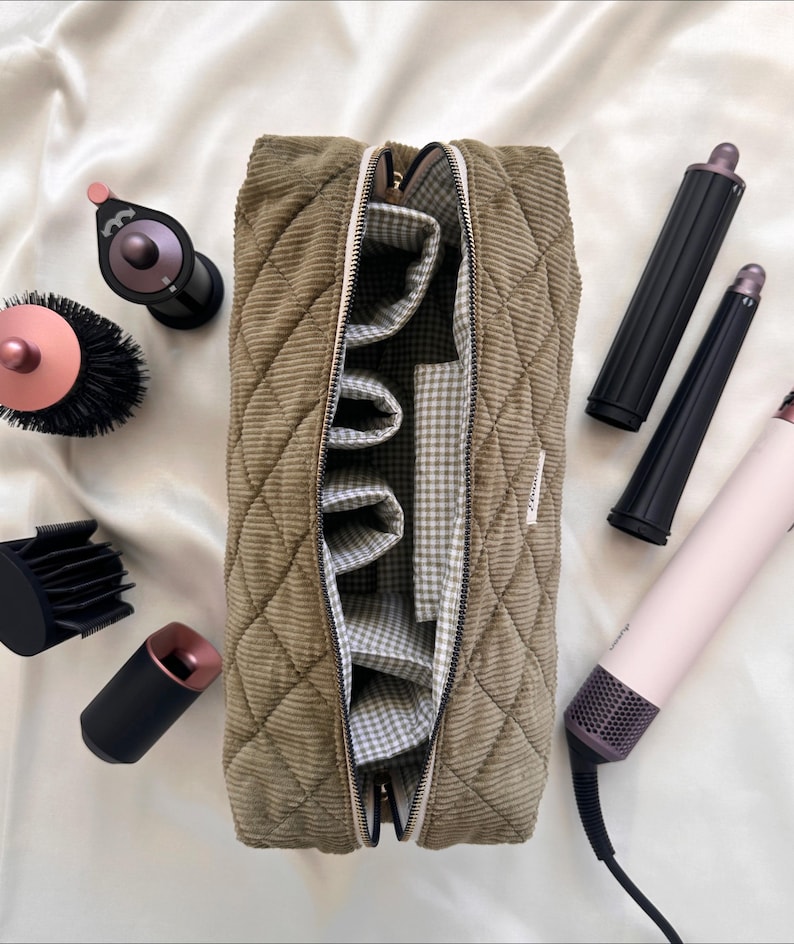May include: A corduroy cosmetic bag in olive green, open to reveal a gingham lining and interior pockets. Various hair styling tools, including a hair dryer, curling wands, and brushes, are arranged around the bag on a white surface.