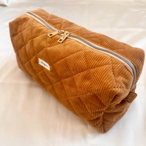 May include: A rectangular, quilted corduroy bag in a warm brown hue. The bag features a silver zipper with gold-colored pulls and a small white label with text. The corduroy has a ribbed texture, and the bag is diamond-quilted.
