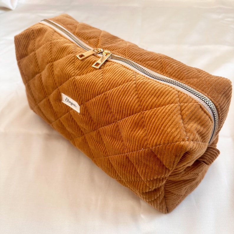 May include: A rectangular, quilted corduroy bag in a warm brown hue. The bag features a silver zipper with gold-colored pulls and a small white label with text. The corduroy has a ribbed texture, and the bag is diamond-quilted.