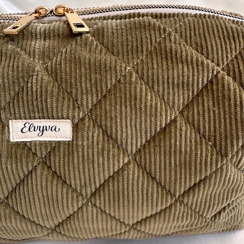 May include: Close-up of a quilted corduroy pouch in a muted olive green color. The pouch features a diamond pattern and a gold zipper. A small label with the name "Elvyva" is sewn on the front.