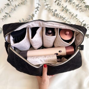 May include: Black corduroy travel organizer bag with multiple compartments. The interior features white fabric with black polka dots, holding hair styling tools. A hair dryer and brush are visible. The bag is open, revealing the contents.