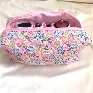 Può includere: Un beauty case trapuntato floreale con cerniera e interno rosa. La borsa presenta un motivo di fiori rosa, blu e bianchi su sfondo crema. L'interno è in tessuto a righe rosa e bianco e ha scomparti per trucchi. Il marchio "Elvyva" è visibile.