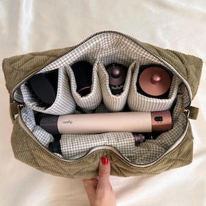 Pode incluir: Uma bolsa de viagem acolchoada de veludo cotelê verde-oliva com fecho de correr, aberta para revelar compartimentos com ferramentas de styling de cabelo. O interior apresenta um padrão xadrez vichy. Uma mão com verniz vermelho segura a bolsa.