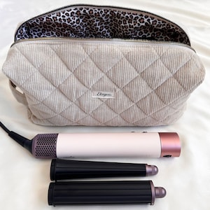 Peut inclure: Un sac &agrave; cosm&eacute;tiques en velours c&ocirc;tel&eacute; beige matelass&eacute;, doubl&eacute; d'un imprim&eacute; l&eacute;opard, ouvert pour r&eacute;v&eacute;ler l'int&eacute;rieur. Un outil de coiffure blanc et or rose et deux accessoires de coiffure noirs sont plac&eacute;s devant le sac. Le sac porte le texte "Elvyva".