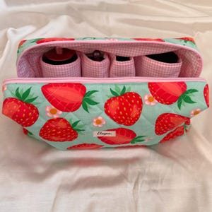 May include: A quilted cosmetic bag with a strawberry pattern. The bag is light blue with large red strawberries and green leaves. The interior has pink gingham lining and small compartments. The bag is open, revealing the interior.