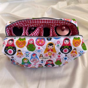 Pode incluir: Uma bolsa de cosméticos acolchoada branca com zíper, com um padrão de bonecas Matryoshka coloridas. O interior tem forro xadrez vermelho e branco e compartimentos para pincéis de maquiagem e outros itens.