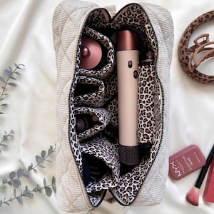 Peut inclure: Un sac &agrave; cosm&eacute;tiques beige matelass&eacute; avec un int&eacute;rieur &agrave; imprim&eacute; l&eacute;opard, ouvert pour r&eacute;v&eacute;ler des outils de coiffure. Un s&egrave;che-cheveux, un fer &agrave; friser et une brosse sont visibles. Une pince &agrave; cheveux et du maquillage sont sur le c&ocirc;t&eacute;.