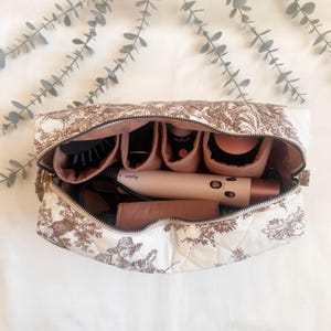 May include: A quilted toiletry bag with a brown and white pastoral design, open to reveal various hair styling tools. The bag features multiple compartments and a zipper closure. A Dyson hair dryer is visible inside.