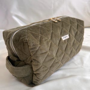May include: Olive green corduroy cosmetic bag with a quilted diamond pattern. The rectangular bag features a gold zipper and a small white label with the name "Ebaya". The bag is designed for storing makeup and toiletries.