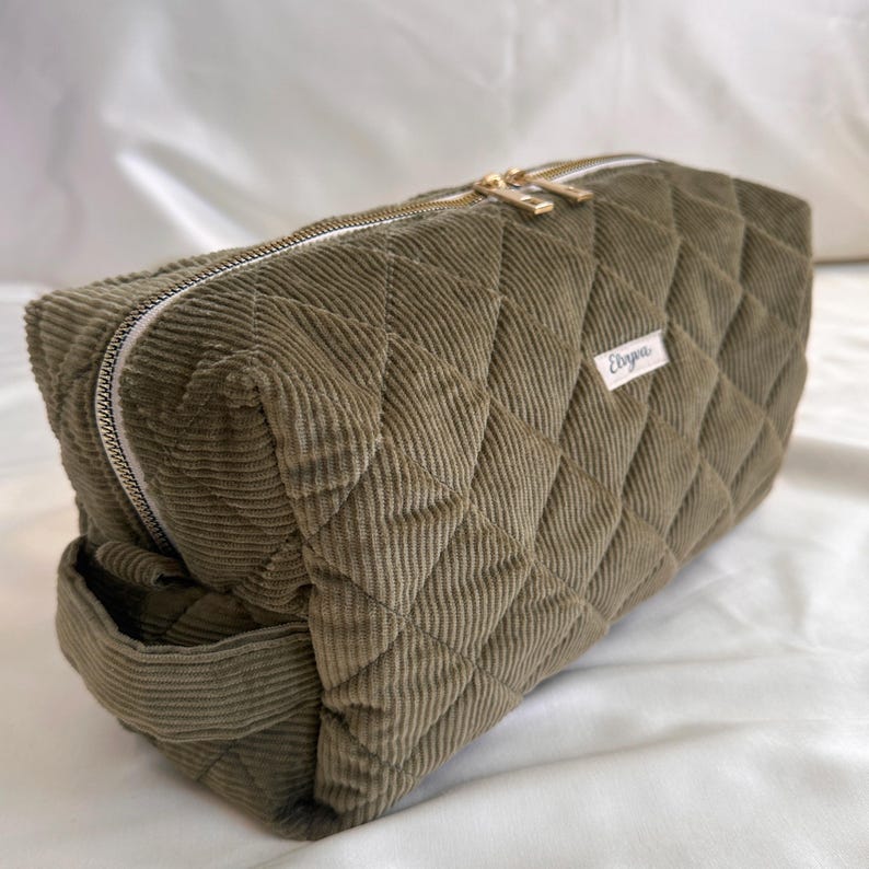 May include: Olive green corduroy cosmetic bag with a quilted diamond pattern. The rectangular bag features a gold zipper and a small white label with the name "Ebaya". The bag is designed for storing makeup and toiletries.
