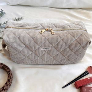 Peut inclure: Une trousse de maquillage en velours c&ocirc;tel&eacute; beige clair matelass&eacute;, avec une fermeture &eacute;clair dor&eacute;e et une petite &eacute;tiquette &laquo; Eenvva &raquo;. Le sac a une forme rectangulaire et une poign&eacute;e sur un c&ocirc;t&eacute;. Le sac est sur une surface blanche.