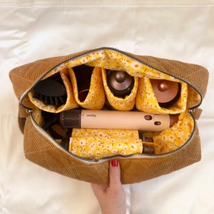 May include: A brown corduroy travel bag with a zipper, open to reveal compartments lined with yellow floral fabric. The bag contains hair styling tools, including a Dyson hair dryer.