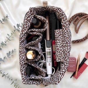 Peut inclure: Un sac &agrave; cosm&eacute;tiques &agrave; imprim&eacute; l&eacute;opard contenant divers produits de beaut&eacute;. On y trouve un fer &agrave; friser, un lisseur, des flacons et du maquillage. Une pince &agrave; cheveux marron et des pinceaux de maquillage sont &agrave; c&ocirc;t&eacute;.