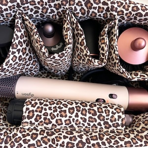 Peut inclure: Un sac de cosm&eacute;tiques &agrave; imprim&eacute; l&eacute;opard avec plusieurs compartiments, contenant divers outils de coiffure. Le sac contient un s&egrave;che-cheveux Dyson, une brosse ronde et d'autres accessoires de coiffure. L'int&eacute;rieur et l'ext&eacute;rieur du sac pr&eacute;sentent un imprim&eacute; animalier marron et beige.