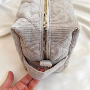 Peut inclure: Un sac &agrave; cosm&eacute;tiques en velours c&ocirc;tel&eacute; beige clair avec un motif matelass&eacute; en losanges. Le sac est dot&eacute; d'une fermeture &eacute;clair dor&eacute;e au centre et d'une petite boucle sur le c&ocirc;t&eacute;. Le sac est en tissu doux et textur&eacute;.