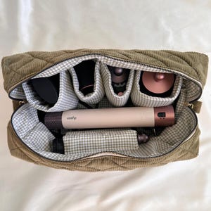 May include: Olive green quilted corduroy cosmetic bag with a zipper closure. The interior features multiple compartments lined with a gray and white gingham pattern. The bag contains hair styling tools, including a Dyson hair styler.